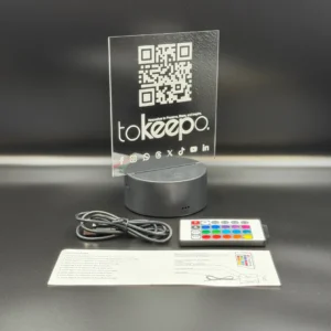 LED Acrylic Desk Lamp by tokeepo with a glowing acrylic plaque featuring engraved designs, including a QR code and logo, illuminated on a laser engraved transparent acrylic. USB cable, remote control, and manual are also in the picture.