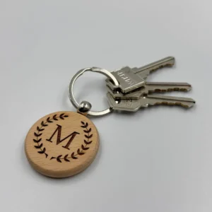 A round wooden keychain with the monogram "M" engraved on its surface, attached to a silver keyring, placed on a white background.