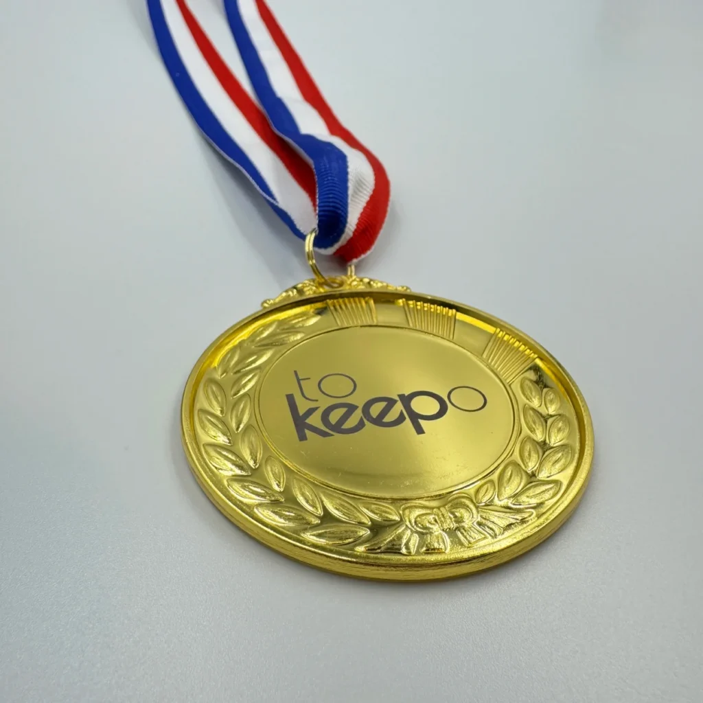 A gold medal with neck ribbon featuring a laurel wreath design, laser marked with the "tokeepo" logo in the center, hanging from a red, white, and blue ribbon.