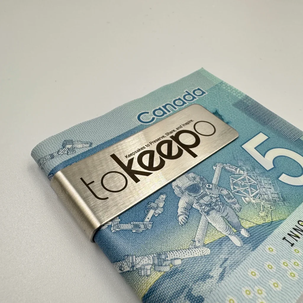 A stainless steel money clip engraved with the "tokeepo" logo, securing folded Canadian five-dollar bills on a white surface.