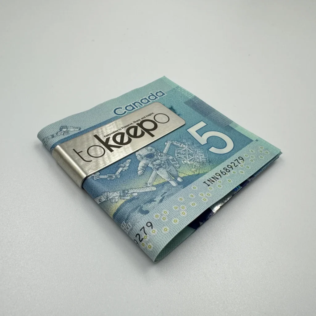A stainless steel money clip engraved with the "tokeepo" logo, securing folded Canadian five-dollar bills on a white surface.