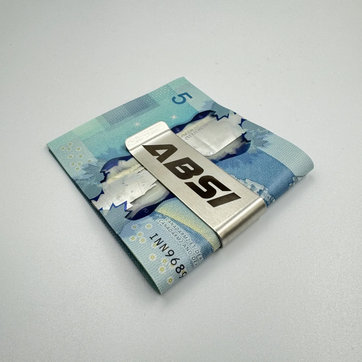 A stainless steel money clip engraved with the "tokeepo" logo, securing folded Canadian five-dollar bills on a white surface.