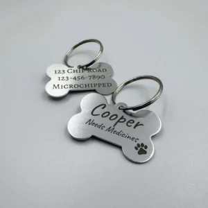 Two stainless steel bone-shaped pet tags, one engraved with "Cooper Needs Medicines" and the other displaying an address, phone number, and microchip information.