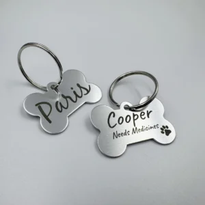 Two stainless steel bone-shaped pet tags with engraved names, "Paris" and "Cooper," attached to keyrings.