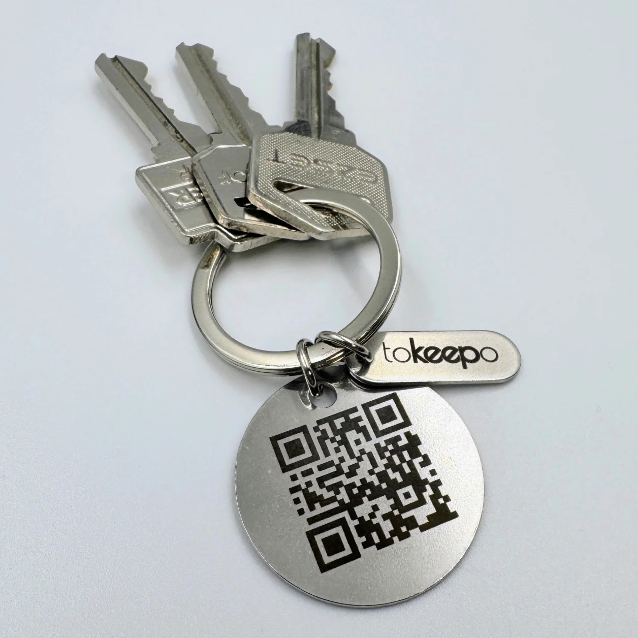 A stainless steel keychain with a circular marked pendant and a small rectangular tag, both featuring the "tokeepo" logo.