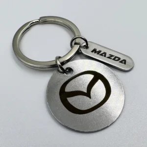 A stainless steel keychain with a circular marked pendant and a small rectangular tag, both featuring the "Mazda" logo.