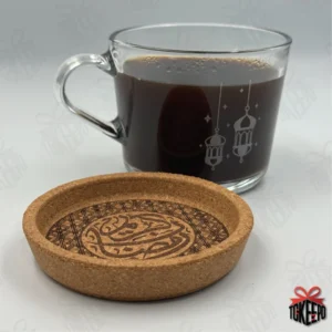 Cork coaster laser engraved with Arabic calligraphy and geometric border, shown beside a glass coffee mug.
