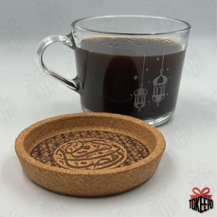 Cork coaster laser engraved with Arabic calligraphy and geometric border, shown beside a glass coffee mug.