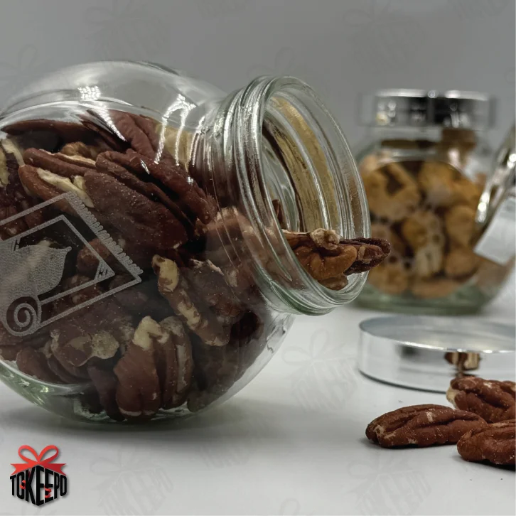 Open engraved Salah reward jar tipped on its side with pecans spilling out, with other jars blurred in the background.